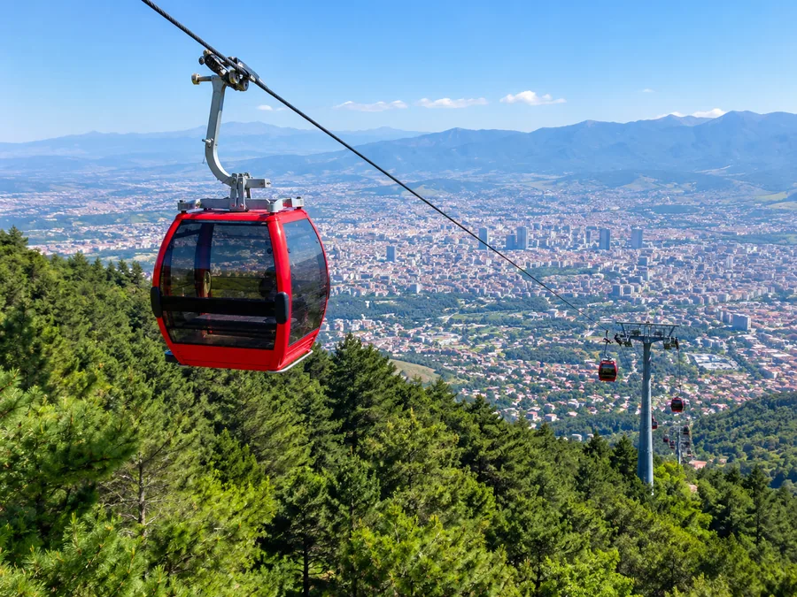 Një kabinë e kuqe e Dajti Ekspres që ngjitet mbi pyllin me pisha, me Tiranën poshtë në luginë